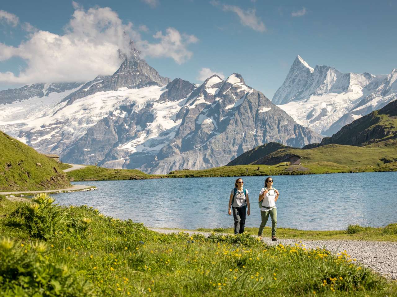 Wandern Bachalpsee Zwei Personen wandern auf einem Pfad, mit dem Bachalpsee und den Bergen im Hintergrund an einem sonnigen Sommertag.