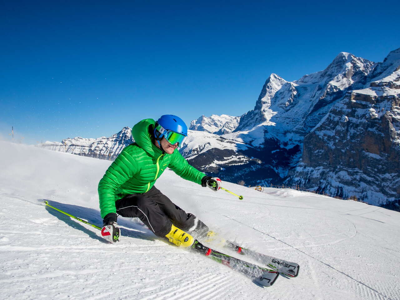 Skifahrer ziehen ihre Kurven im Skigebiet Mürren-Schilthorn. Das Dreigestirn Eiger, Mönch und Jungfrau ist aus der Ferne zu sehen.