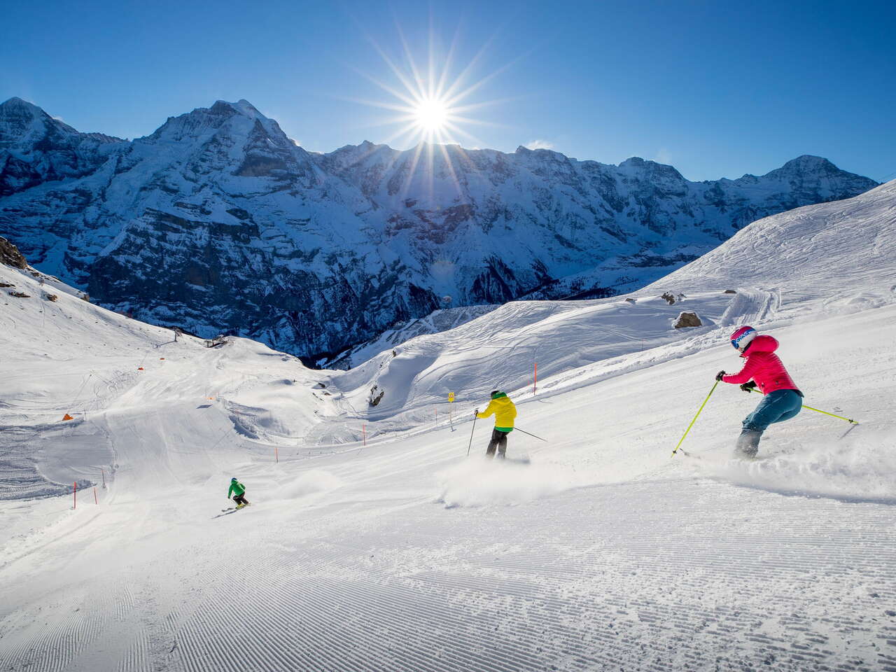 Skifahrer ziehen ihre Kurven im Skigebiet Mürren-Schilthorn. Das Dreigestirn Eiger, Mönch und Jungfrau ist aus der Ferne zu sehen.